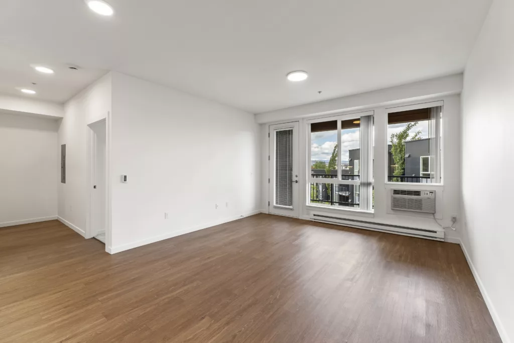 An empty room with wooden floors, white walls, and large windows leading to a balcony with an outdoor view.