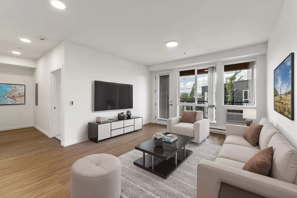 Modern living room with a large TV, beige sofa set, round ottoman, and a wooden coffee table on a light rug, leading to a balcony.