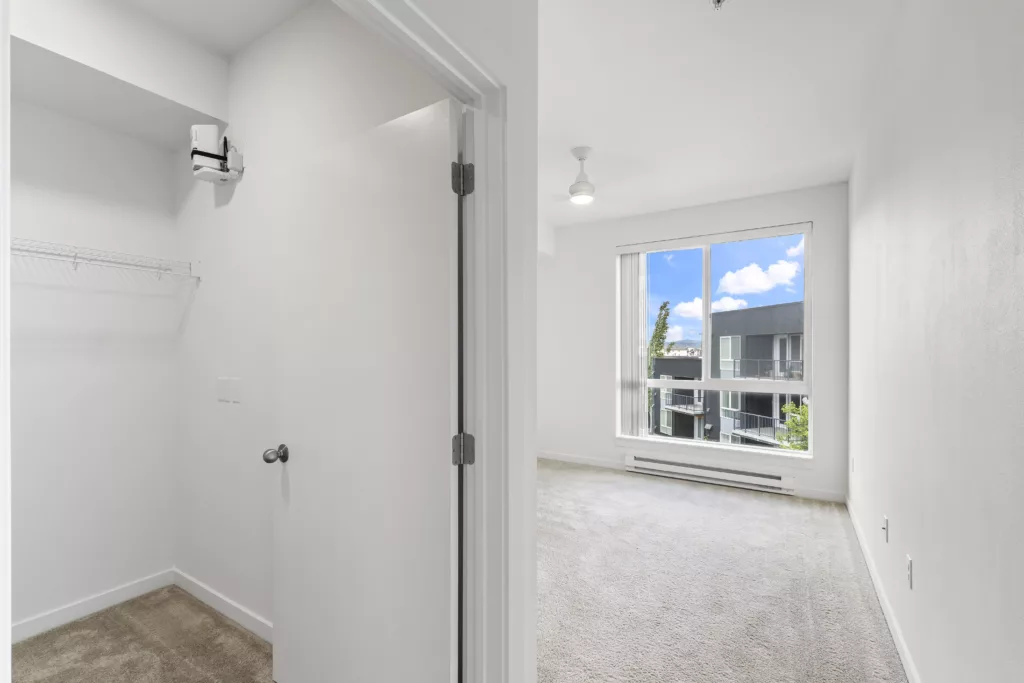 Modern, empty apartment room with a large window, carpeted floor, and an open closet space on the left.