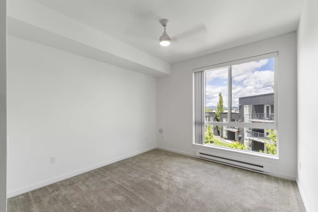 Empty room with gray carpeting, white walls, a ceiling fan, and a large window overlooking a view of neighboring buildings.