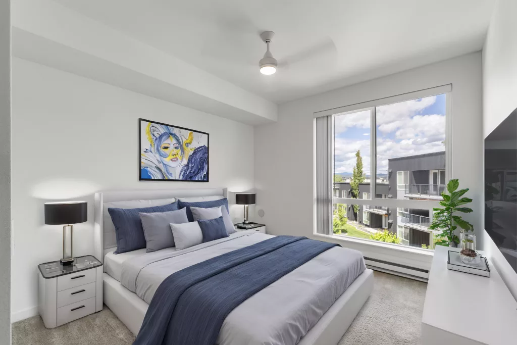 A modern bedroom with a neatly made bed, blue accents, large window, artwork on the wall, and a potted plant by the TV.