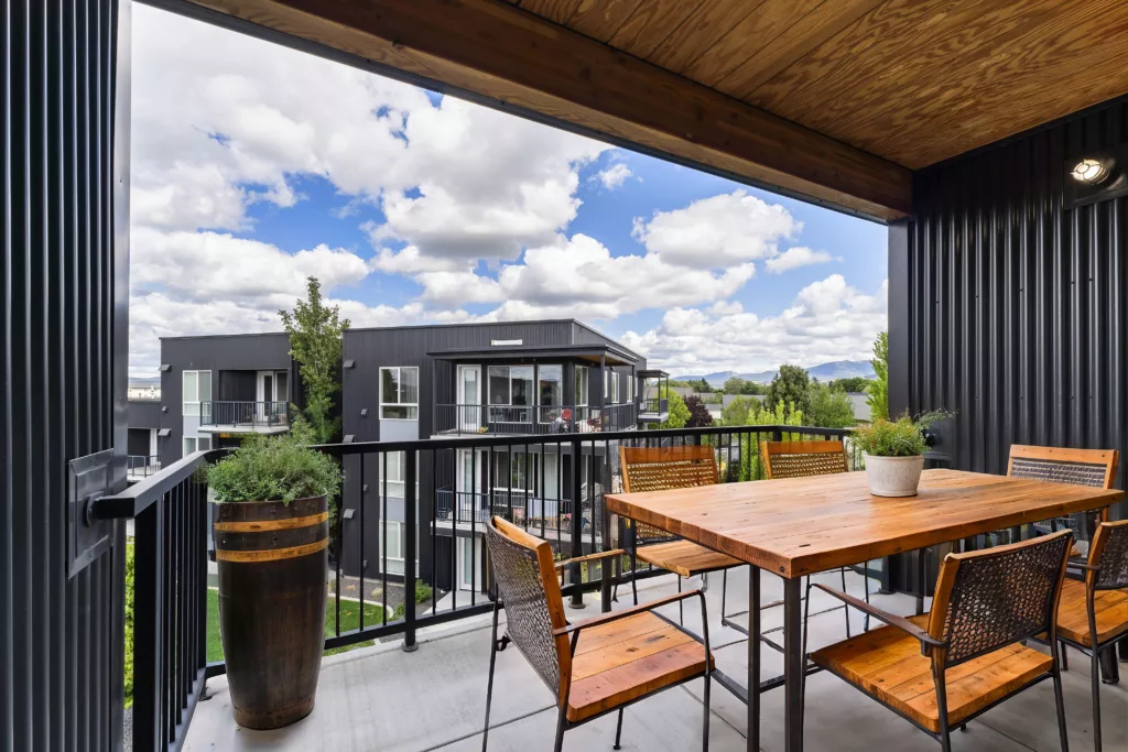 A covered balcony with wooden furniture overlooks modern buildings and a cloudy sky.